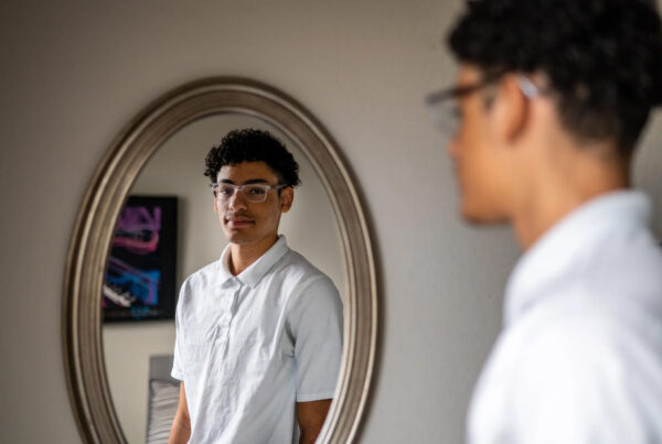 A teenage boy's reflection from the torso up is seen in a oval mirror, the teen himself is seen from the side standing to the right of the photo looking into the mirror. He has dark curly hair and clear-framed glasses and is sporting a white polo. This is Myles Leon.