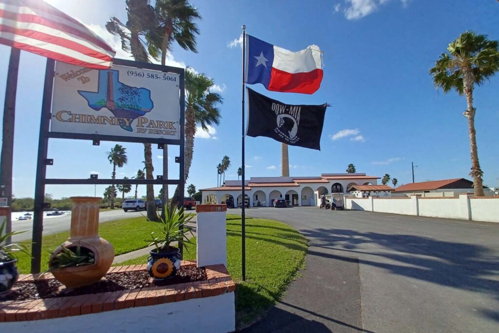 A sign that says "welcome to Chimney Park RV resort" is seen in the foreground next to U.S., Texas and POW flags. A resort building is in the background.