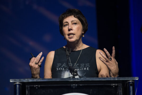 Nicola Fox gestures with both hands while speaking at a podium. She is wearing a sleeveless black shirt that says "Parker Solar Probe" on it.