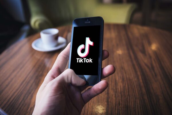 A close-up of a hand holding an iPhone displaying the TikTok app logo on its screen. A table with a cup of coffee are in the background.