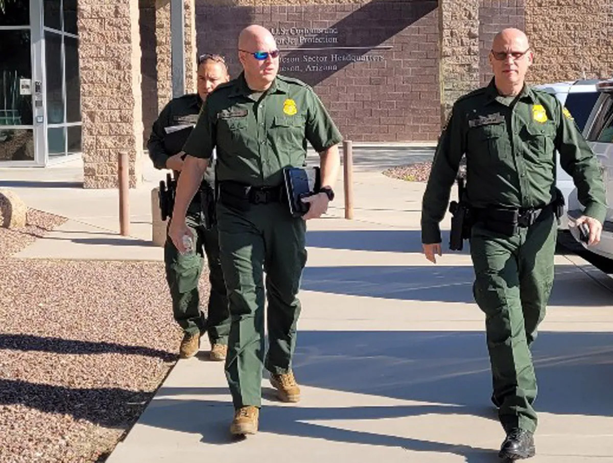 Three border patrol officers are seen walking away from a building, dressed in their green uniforms. At left is Tony Barker, wearing sunglasses and seemingly in mid-sentence talking to the officer to his left.