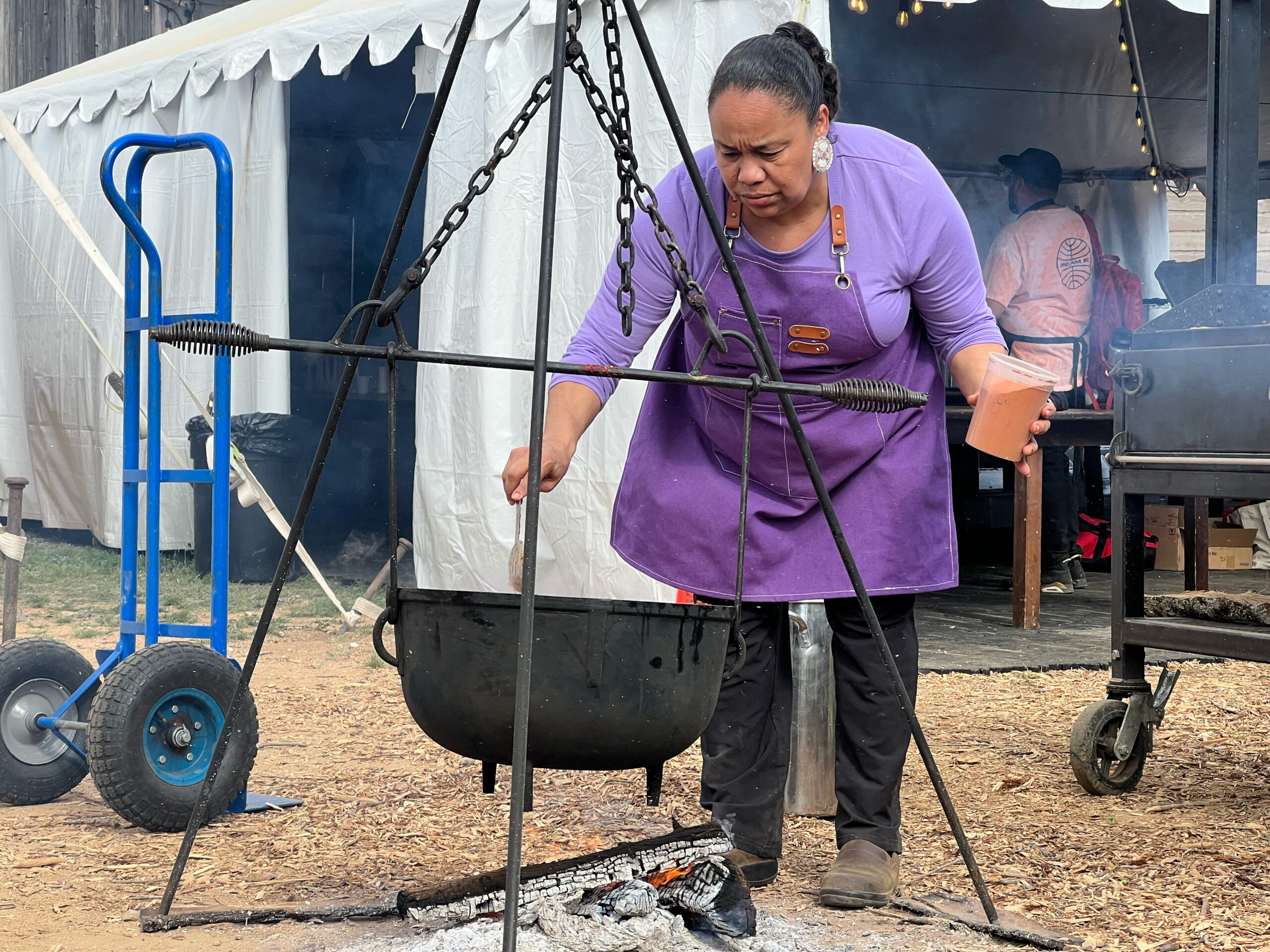 A woman stirs a cast iron put suspended over an open flame. This is chef Crystal Wahpepah.