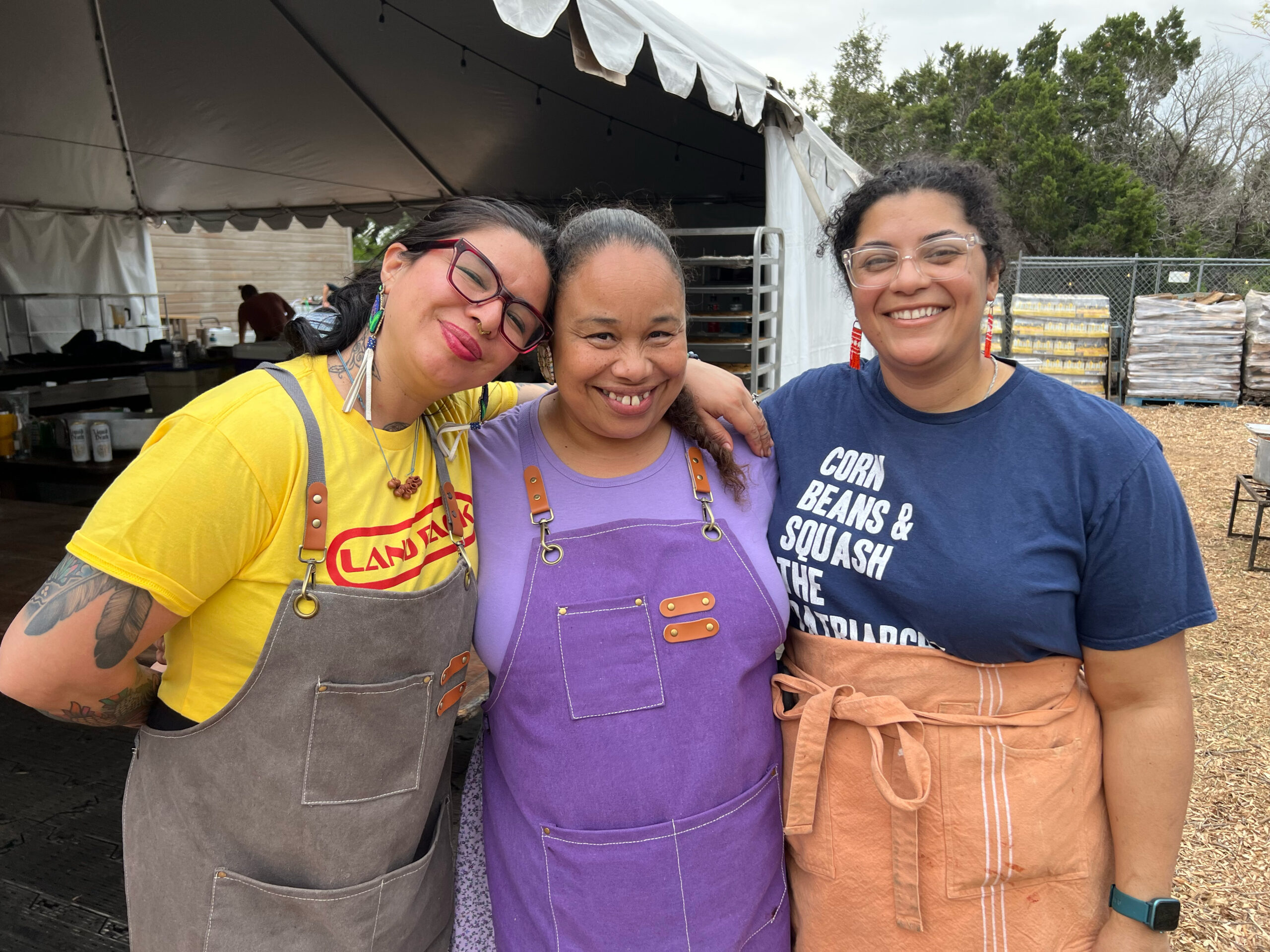 Three people smile for the camera, their arms on each others shoulders in an embrace. These are the chefs features at the 2023 Potluck dinner: Sewa Yuli, Crystal Wahpepah and Britt Reed.