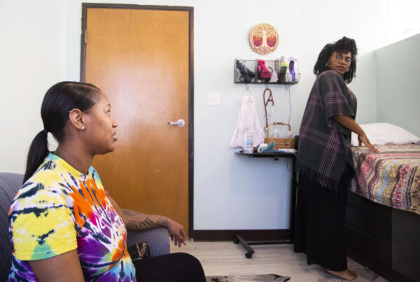 A seated woman speaks with another woman across the room from her who is turning back to answer her. The seated woman is Cherish Sims and she is speaking to her nurse midwife., Krystal Brown. They appear to be in a doctor's office.