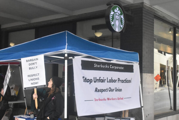 A worker is seen picketing under a tarp outside a Starbucks locations, the sign of which can be seen above the tarp. The worker holds a sign reading in all caps "Bargain, don't bully" and "respect unions now!" Another sign attached to the tarp reads "Stop unfair labor practices! Respect our union. Starbucks Workers United."