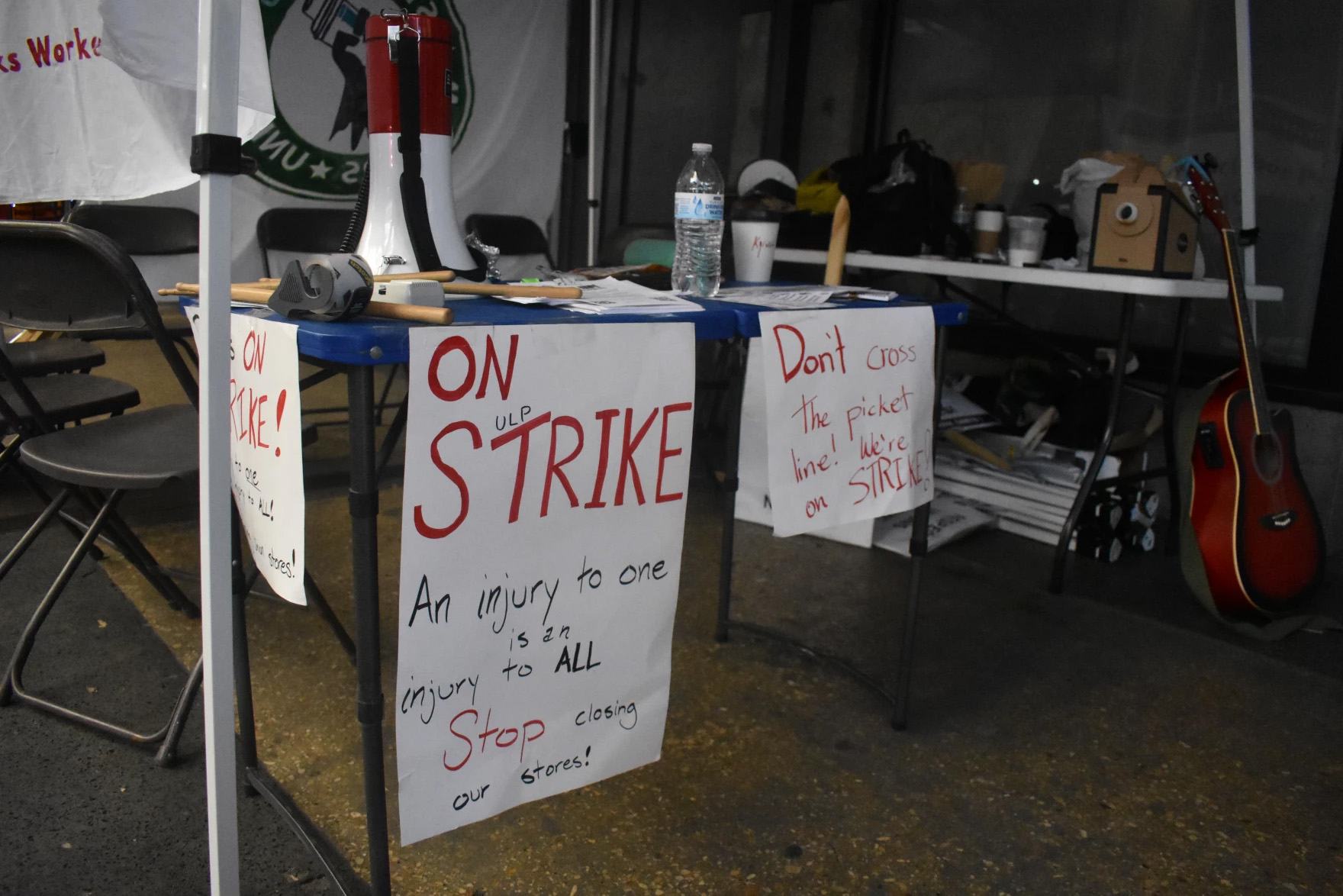 A table is seen at a Starbucks Workers United picket. On the table is a megaphone and two signs are draped on it. One reads 