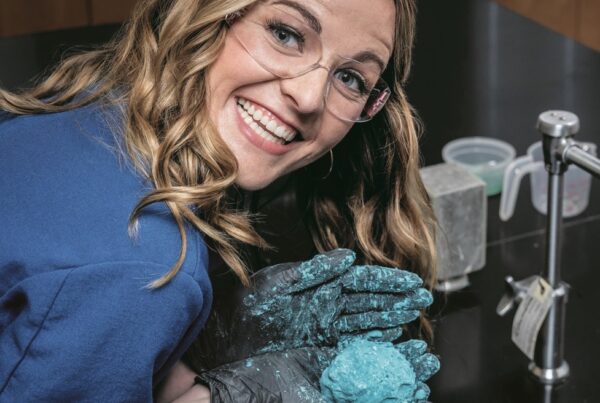 A smiling woman wearing eye protection glasses holds a messy blue shiny ball in her gloved hands.