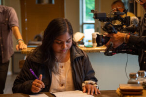 A student focuses on writing at a desk while someone behind is working with a video camera.