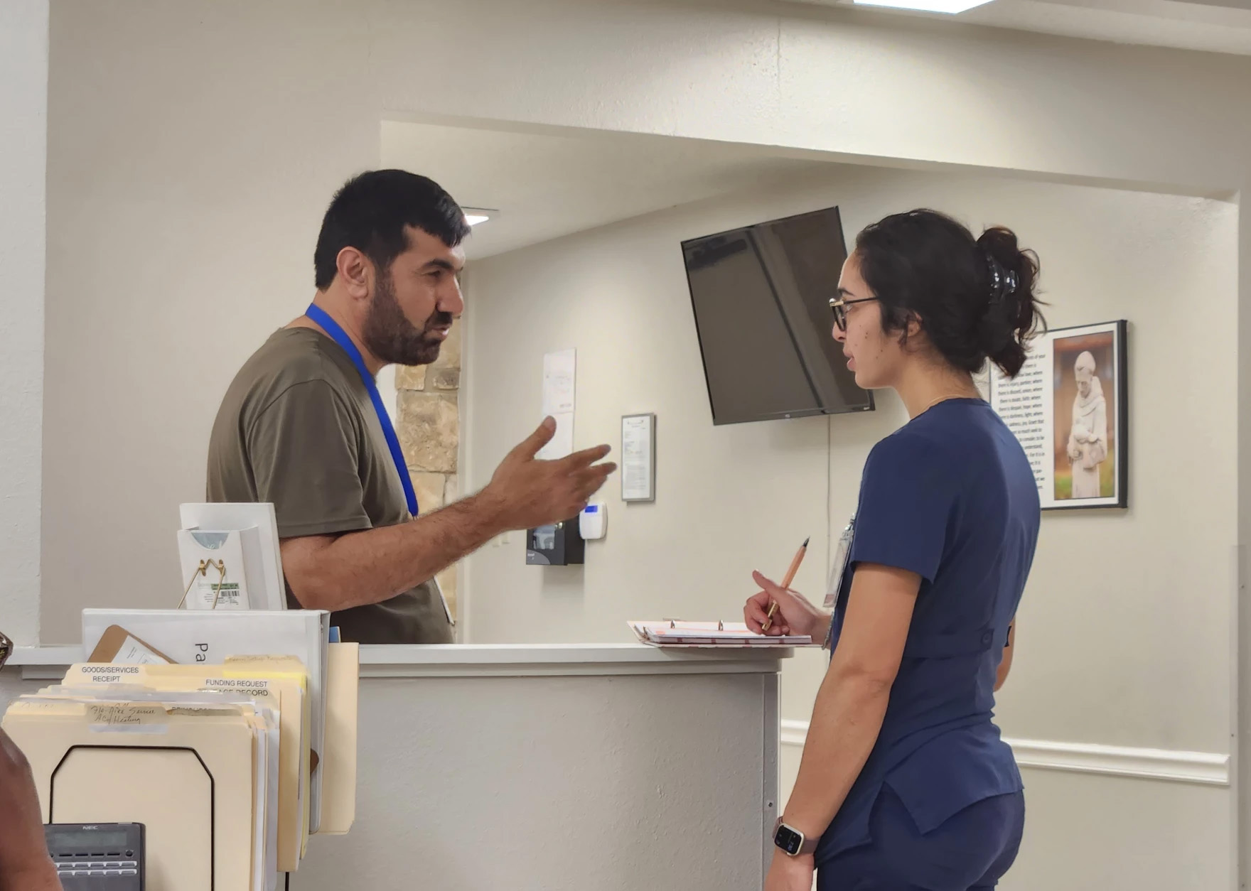 A bearded man in scrubs speaks to a woman, also in scrubs, at the counter at a medical clinic. The man is Chinar Sedeqi.