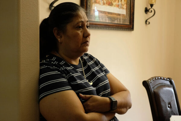 A woman leans against a wall, arms folded, looking off to the side with a concerned look on her face. This is Araceli Palomino.