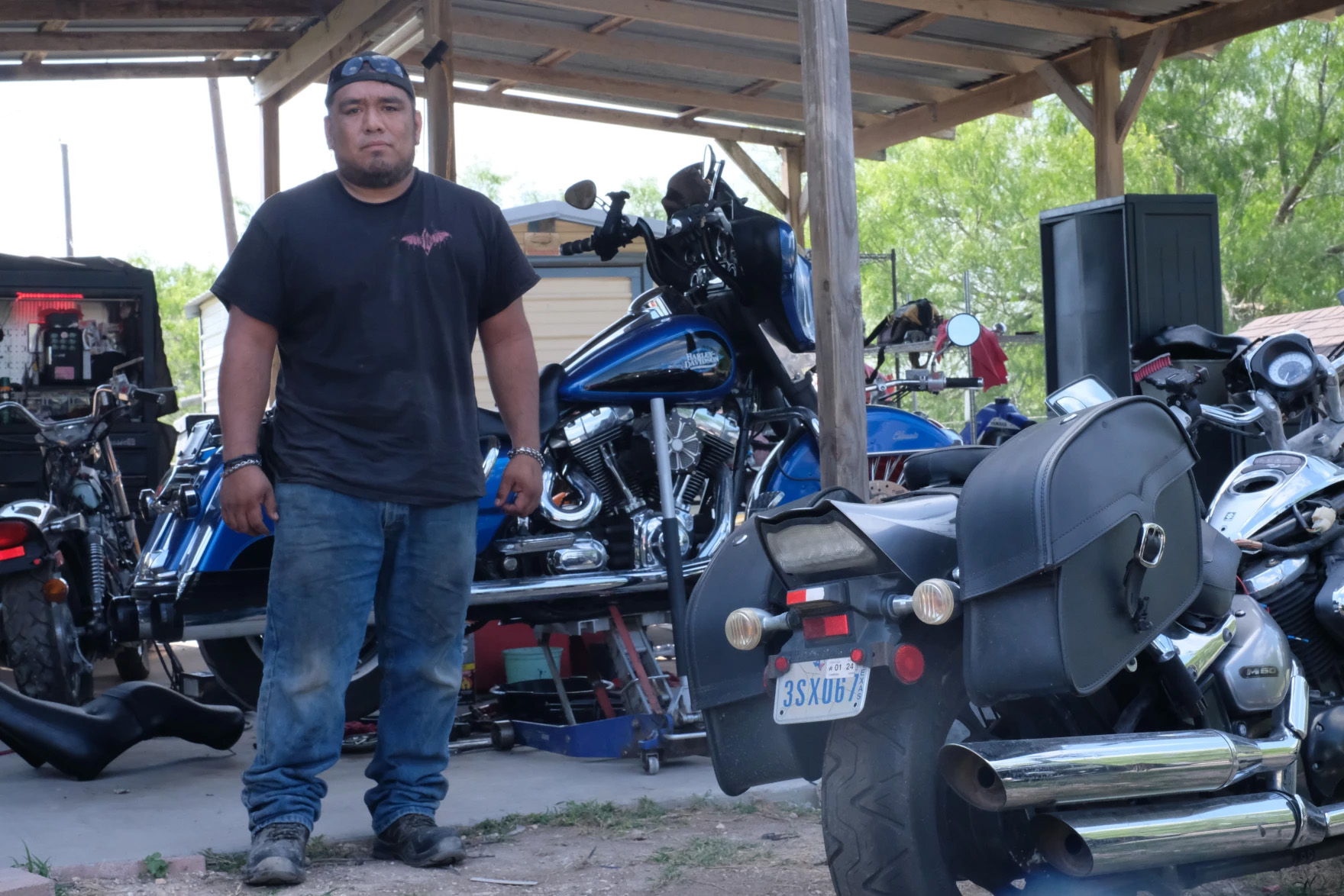 A man stands outside, facing the camera, under an awning. Several motorcycles are seen behind him and in the foreground. He's wearing a t-shirt and blue jeans that show some wear. This is Edgar Flores.