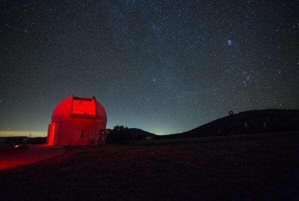 The domed roof over a very large telescope glows red and stars shine in the night sky above a mountainous landscape.