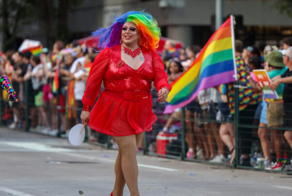 A person dressed in drag – a red dress and heels with a rainbow wig and makeup – walks in a Pride parade. Onlookers are seen in the background with a rainbow Pride flag held by one.