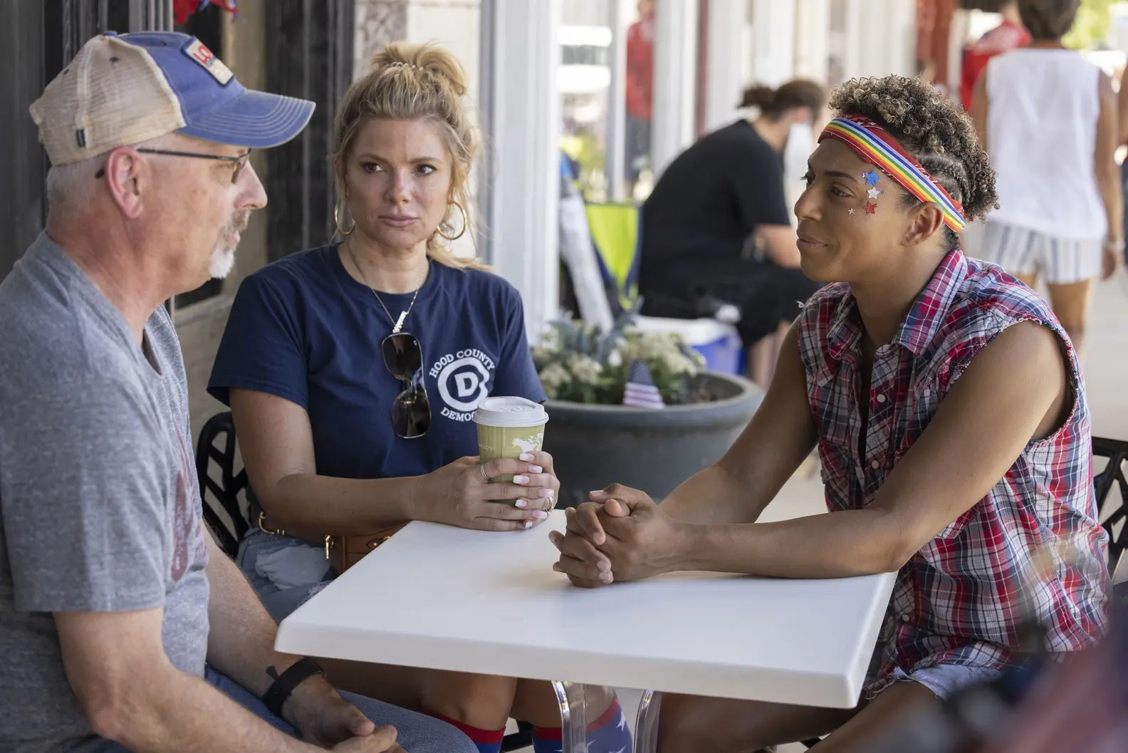 Three people sit at a table in discussion. From right, an older man speaks across the table to Shangela, a drag queen wearing a sleeveless plaid top, rainbow headband and star stickers around their eyes. In the middle a woman looks on.