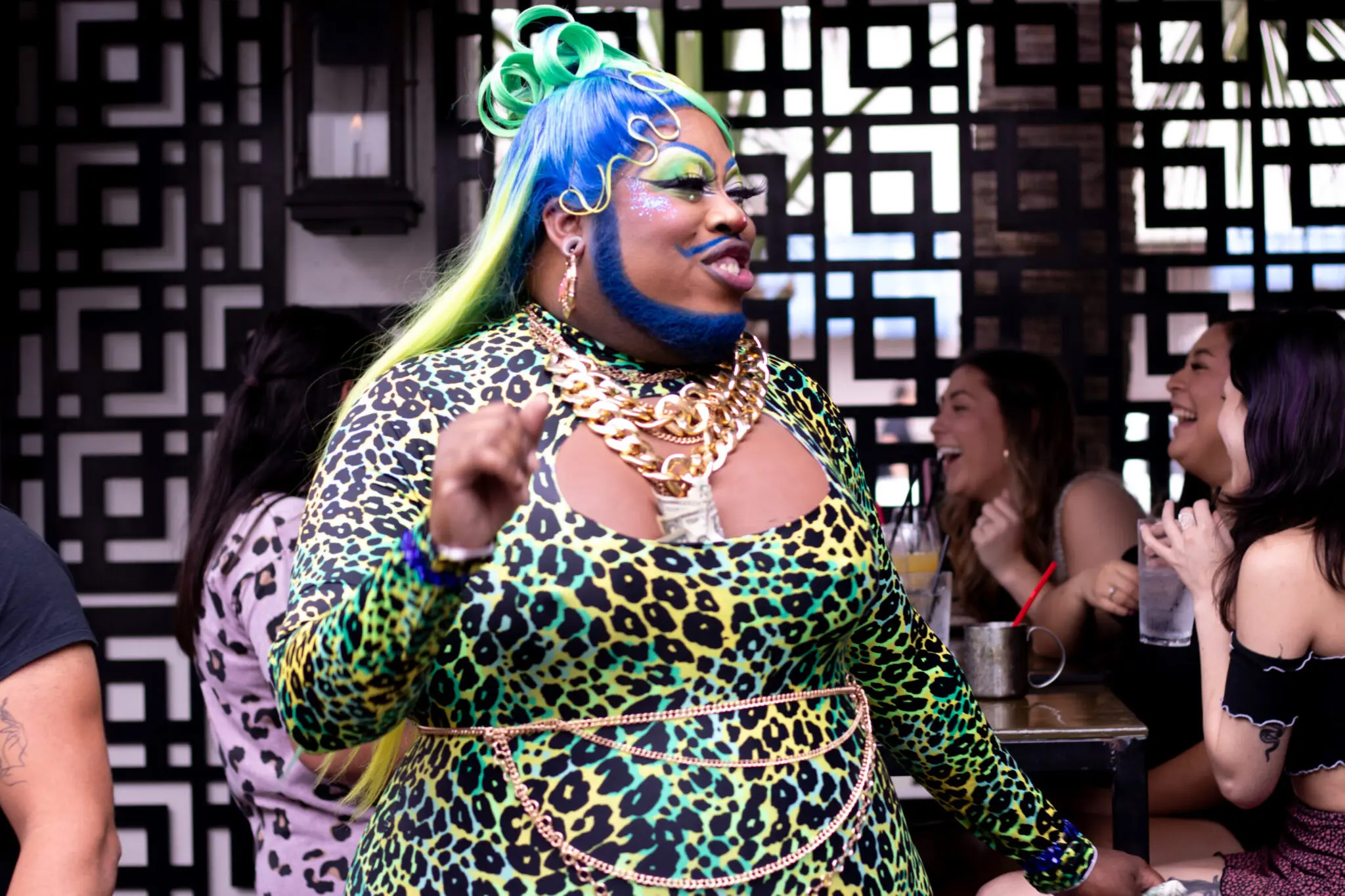 A drag performer dressed in multicolor jaguar print, multicolor hair, and a blue-dyed beard speaks. This is Bearded Beauty Blackberri.