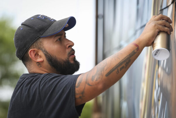A man sprays pain on the side of a building as part of a mural he is working on. This is Juan Velazquez.