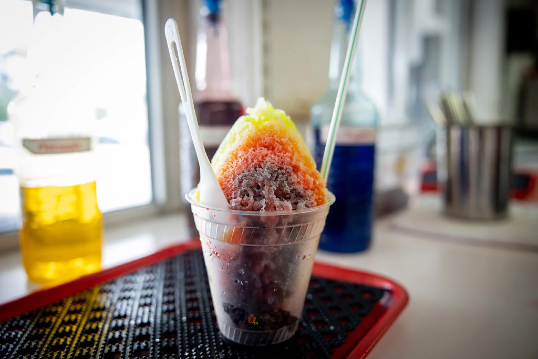 A snow cone is seen with a straw and spoon sticking out of the cup. The shaved ice is yellow, red and purple in color.