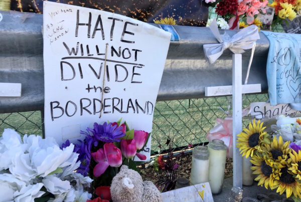 A memorial of flowers, candles and signs honoring the victims of the El Paso Walmart shooting sit in front of a chain-link fence. The most prominent sign reads "hate will not divide the borderland."