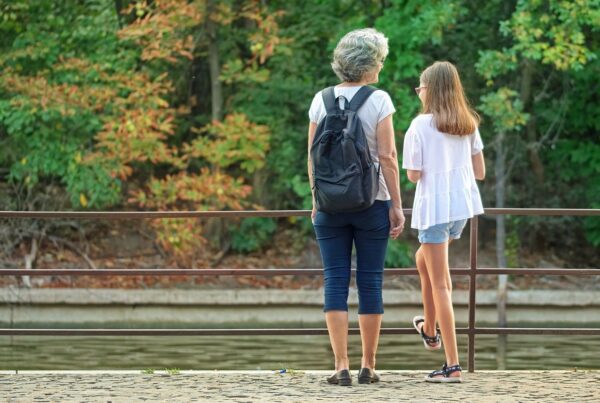An older person with short grey hair stands next to a younger person with long brown hair. The two are looking at a forested area with a waterway and appear to be talking.