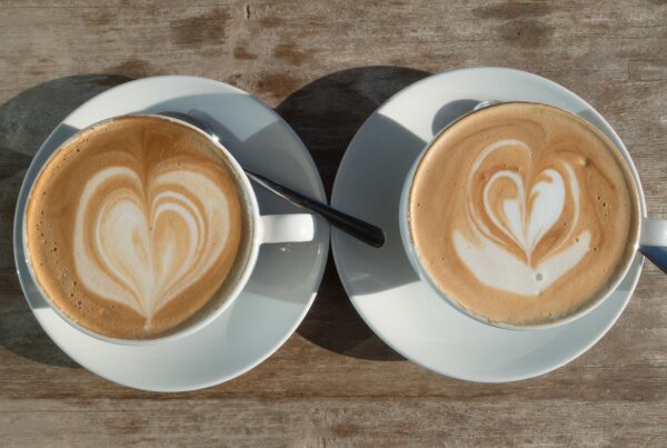 Two cappuccinos sit side by side in white cups on white saucers. The foam in the drinks is in the shape of hearts.