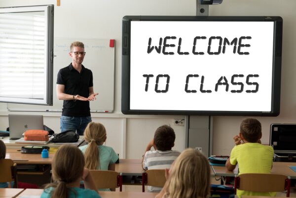 A photo of a teacher standing in front of rows of desks with children looking towards them. A large monitor reads "welcome to class."