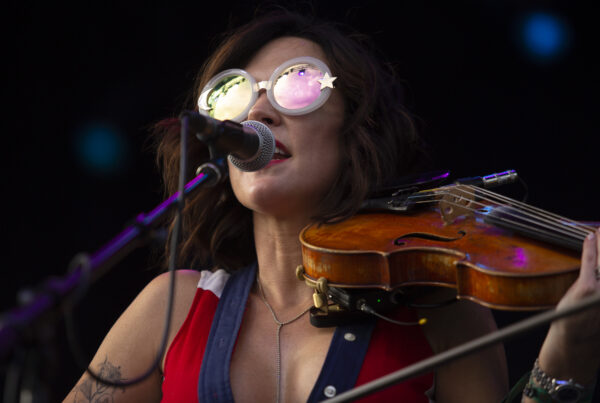 A woman sings into a microphone while playing into a fiddle. She's wearing big reflective sunglasses. This is country music artist Amanda Shires.