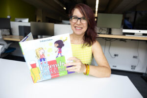 Lalena Fisher wears a yellow dress and smiles for the camera holding her large, square children's book.
