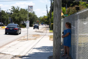 A woman stands in the shade by a powerline pole looking out to the street off to the side on a sunny day.