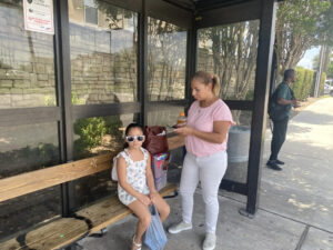 A woman stands under a covered bus stop while her daughter sits, wearing sunglasses, staring at the camera.