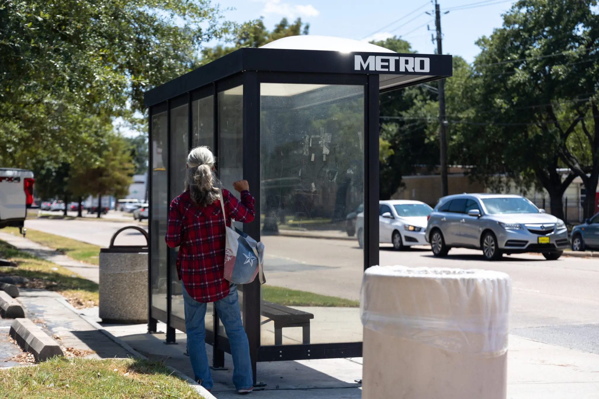 A gray-haired woman waits by the shade of a covered bus stop near a road on a sunny day.