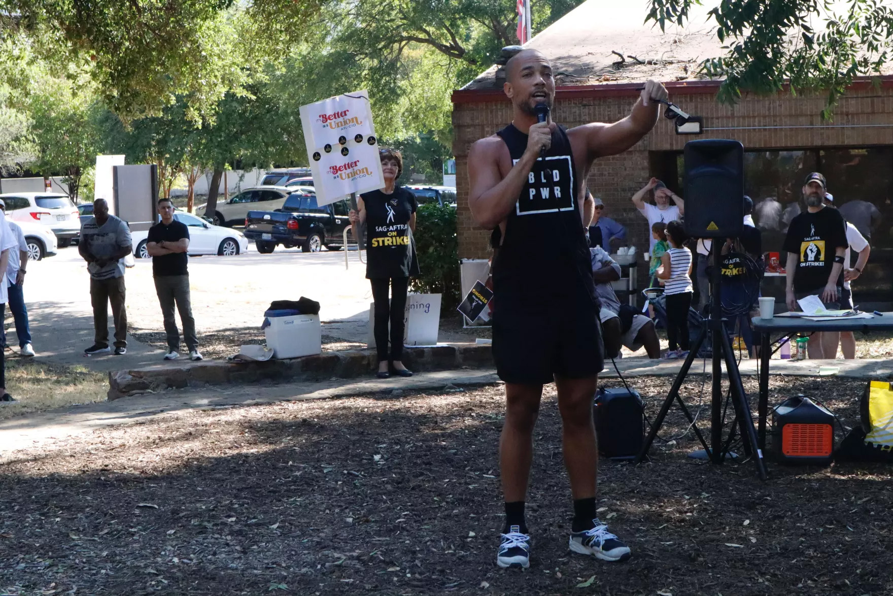 A man holds a microphone speaking at an outside rally. This is actor and union member Kendrick Sampson. He wears a tank top with a stylized spelling of the phrase 