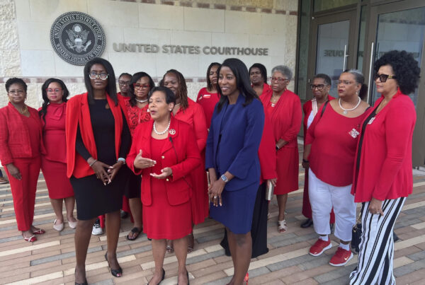 A group of women of varying ages, all dressed professionally and wearing red, except for one wearing blue, gather outside a federal courthouse. One woman in the middle is speaking. They are members of the Delta Sigma Theta Sorority, Inc., a historically Black sorority.