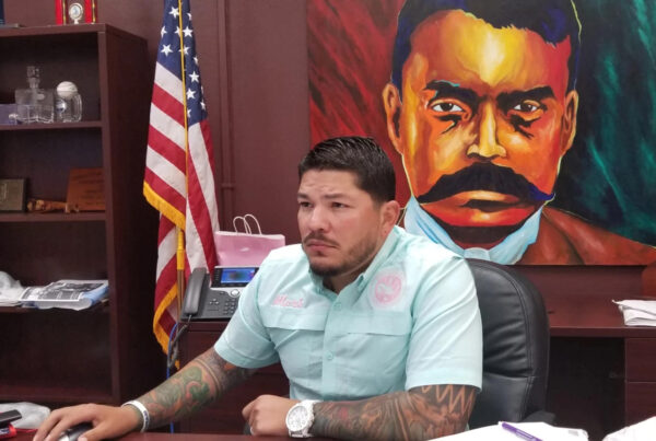 A man sits at a desk in his office, his hand on a mouse and looking at a monitor that's out of the frame. He's wearing a light blue button-up with his name, "Mark," stitched on it and the seal of the Nueces County District Attorney's office. This is Mark Gonzalez. In the background a U.S. flag is seen as well as a painting of Mexican revolutionary Emilio Zapata.