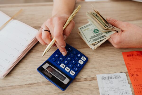 Stock image of a person's hands holding cash in one hand and a pencil in the other while typing on a calculator, suggesting they are working on a budget.