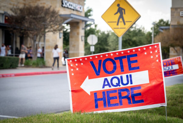 A red sign stuck in the ground says "Vote here/aqui" in blue letters with a white arrow pointing in the direction of a building