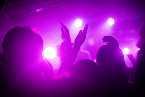 A close-up photos of heads and hands enjoying a concert as a purple light shines in the background.