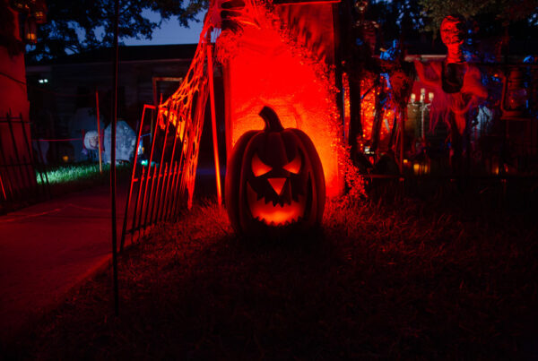 An orange light shines through the face of a plastic jack-o-lantern next to the walkway of a house heavily decorated for Halloween.