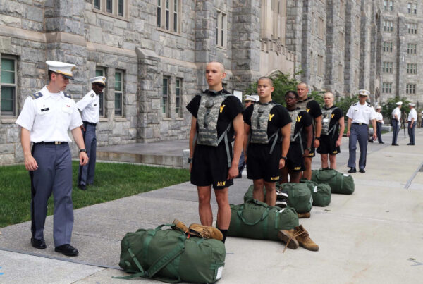 Cadets stand at attention with their belongings in a duffel bag before them as uniformed officers inspect them.