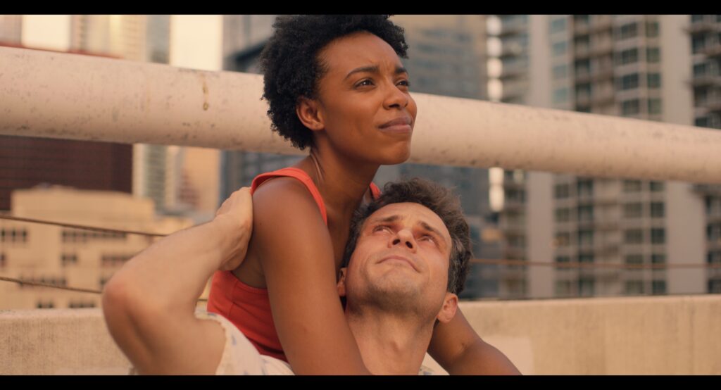 Two people embrace on the roof of a large, concrete building with skyscrapers in the background.