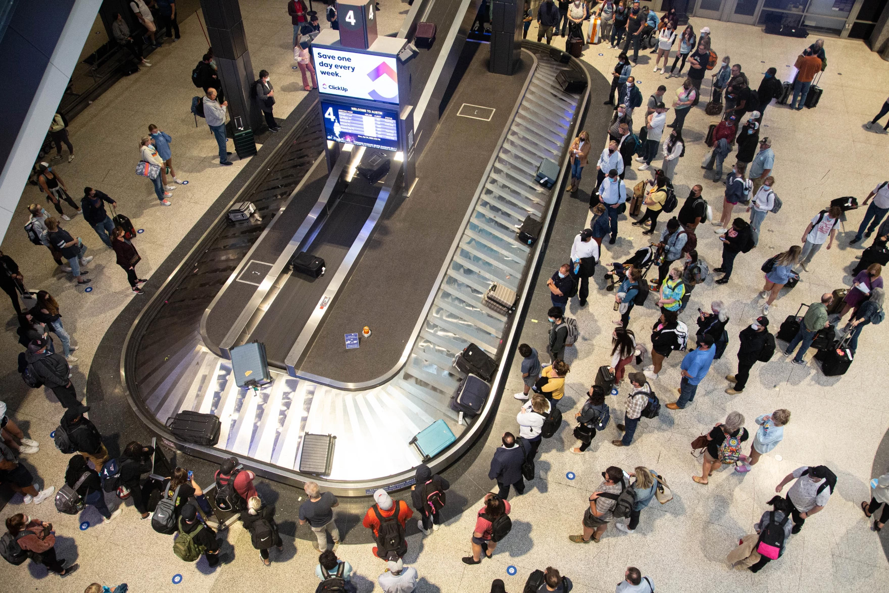 A crowd of people gathered around the baggage claim conveyor belts of an airport are seen from above.