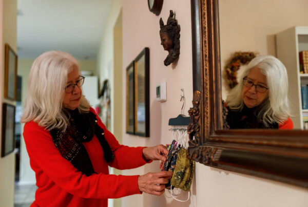 An older woman sorts through several cloth face masks hanging from hooks on a wall in a home. The woman is reflected in a mirror also on the wall. This is Alice Barton.