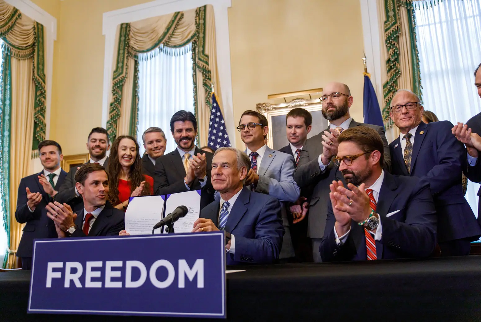 Texas governor Greg Abbott, seated behind a desk, holds up a paper enclosed in a folder he just signed as a group of men and one women in business attire applaud around him. A sign posted at the front of the desk reading 