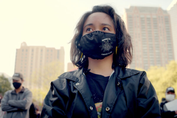 A young woman with shoulder-length brown hair wears a black jacket and a black COVID-19 cloth face mask. The face mask has a floral design on it and reads "kwento ko kewnto natin."