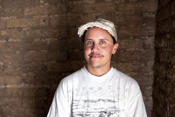 A man in a white shirt and white baseball hats smiles at the camera. He stands in front of an old brick interior wall.
