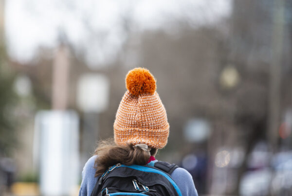 A view from a behind of a woman walking outside wearing a backpack and an orange winter hat