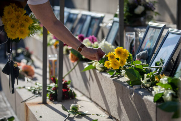 The arm of a person seen out of frame places flowers in front of framed photos of some of the children killed in the Robb Elementary School shooting. More flowers and votive candles are seen among the memorial.