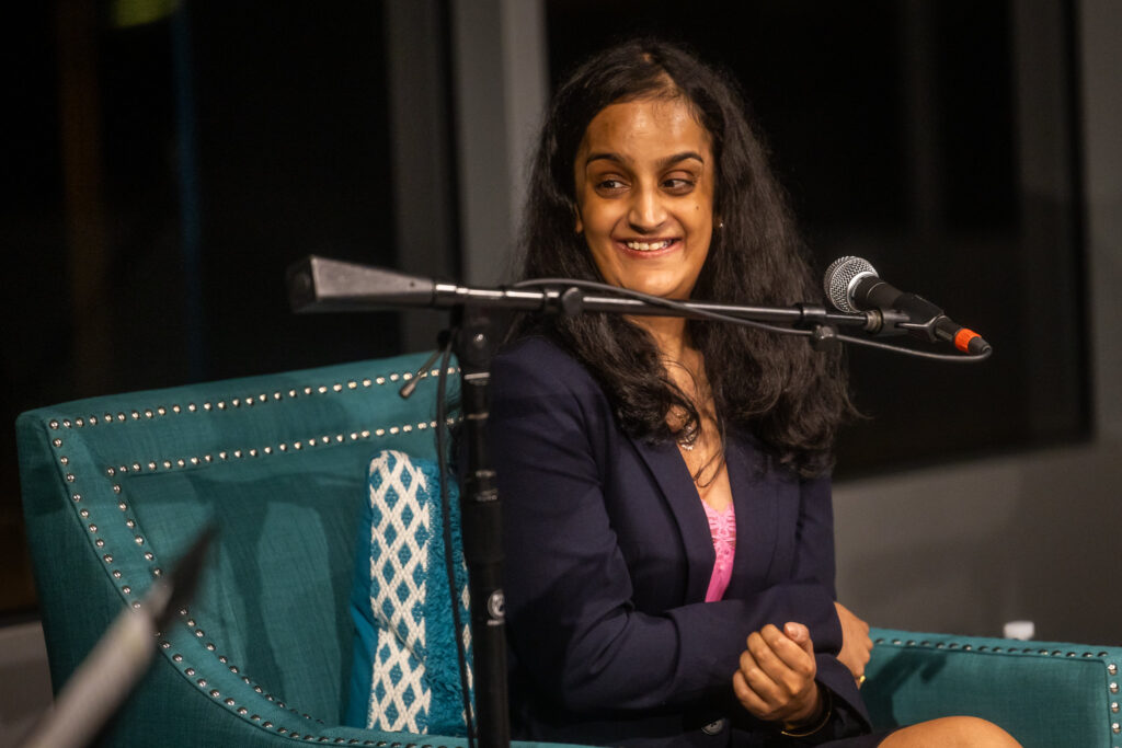 A photo of an Indian American woman with long black hair sitting at a microphone and smiling.