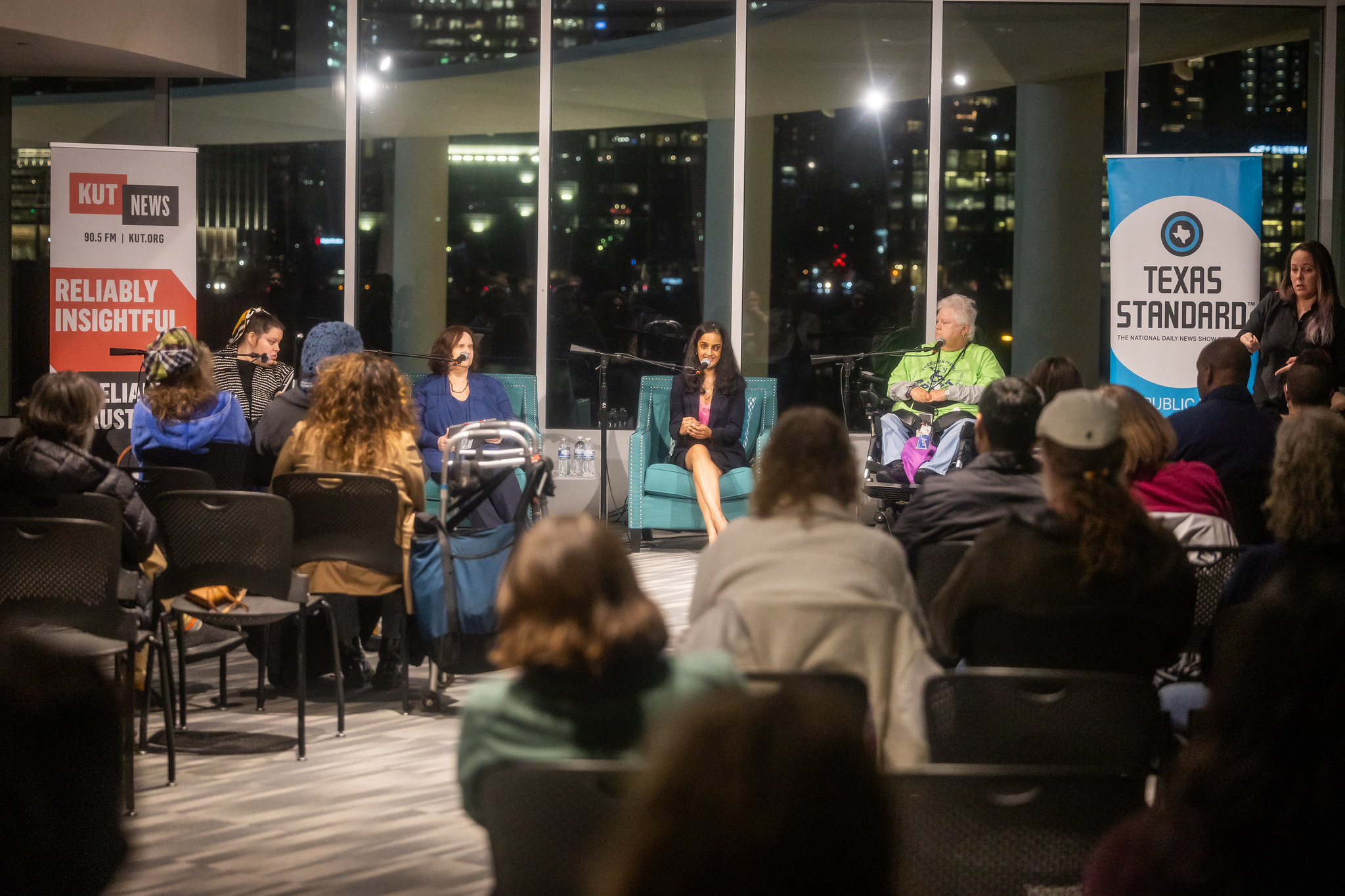 A photo of a room of people sitting in chairs and watching a panel of people at the front of a room. Behind the panelists are large windows that show city buildings lit up at night.