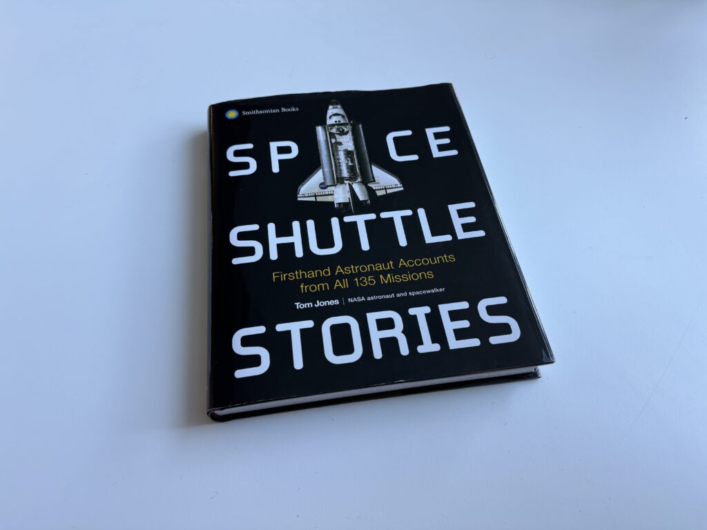 The big, rectangular coffee table book "Space Shuttle Stories" sits on a white table. The cover of the book is black with big white lettering. An image of a space shuttle takes the place of the "a" in space in the title.