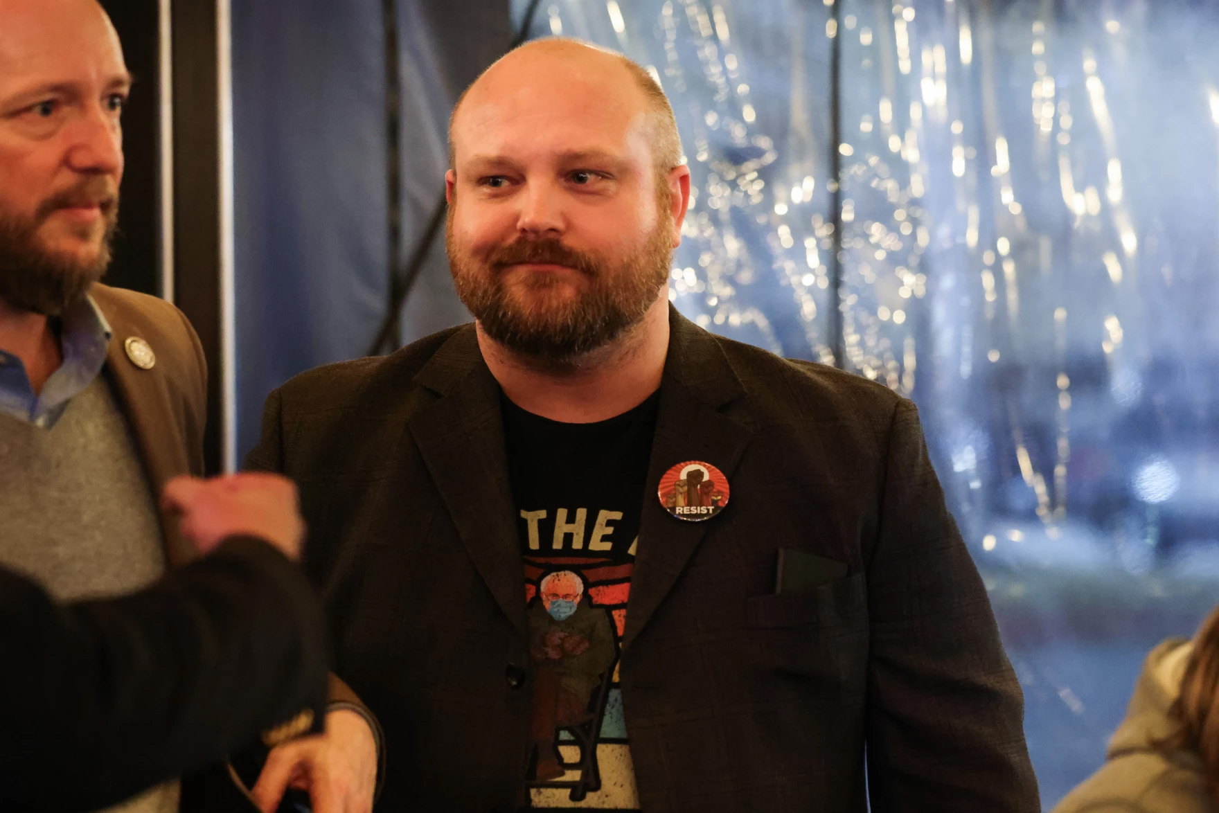 A man grins, looking off to the side. He is bald and sporting a beard, wearing a sport coat over a Bernie Sanders t-shirt and has a button reading 